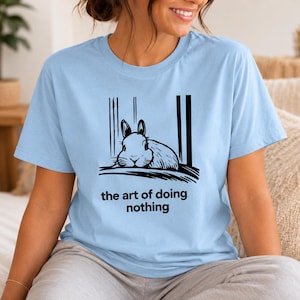 Puede incluir: Una camiseta azul claro con una ilustraci&oacute;n gr&aacute;fica negra de un conejo descansando entre l&iacute;neas verticales. Debajo del conejo, el texto "the art of doing nothing" aparece en una fuente negra, negrita y sin remates centrada en el pecho.