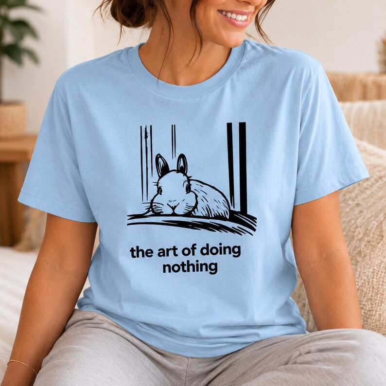 Puede incluir: Una camiseta azul claro con una ilustraci&oacute;n gr&aacute;fica negra de un conejo descansando entre l&iacute;neas verticales. Debajo del conejo, el texto "the art of doing nothing" aparece en una fuente negra, negrita y sin remates centrada en el pecho.