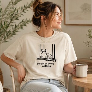 Puede incluir: Una camiseta de manga corta de color crema con una ilustraci&oacute;n gr&aacute;fica negra de un conejo descansando. Debajo del conejo, el texto dice "the art of doing nothing". La camiseta tiene cuello redondo y un corte holgado.