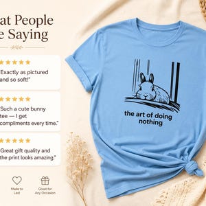 Puede incluir: Una camiseta de manga corta de color azul claro con un gr&aacute;fico negro de un conejo descansando y el texto "the art of doing nothing" impreso debajo. La camiseta se muestra junto a tres rese&ntilde;as de clientes y valoraciones de cinco estrellas.