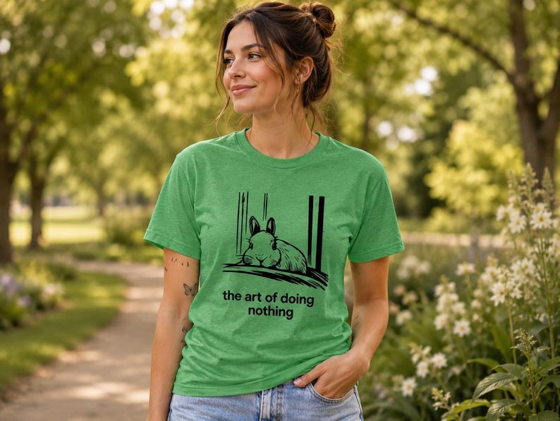Puede incluir: Una camiseta de manga corta en color verde jaspeado con una ilustraci&oacute;n gr&aacute;fica negra de un conejo descansando sobre una superficie. Debajo del conejo, el texto dice, the art of doing nothing. La camiseta tiene cuello redondo y corte est&aacute;ndar.