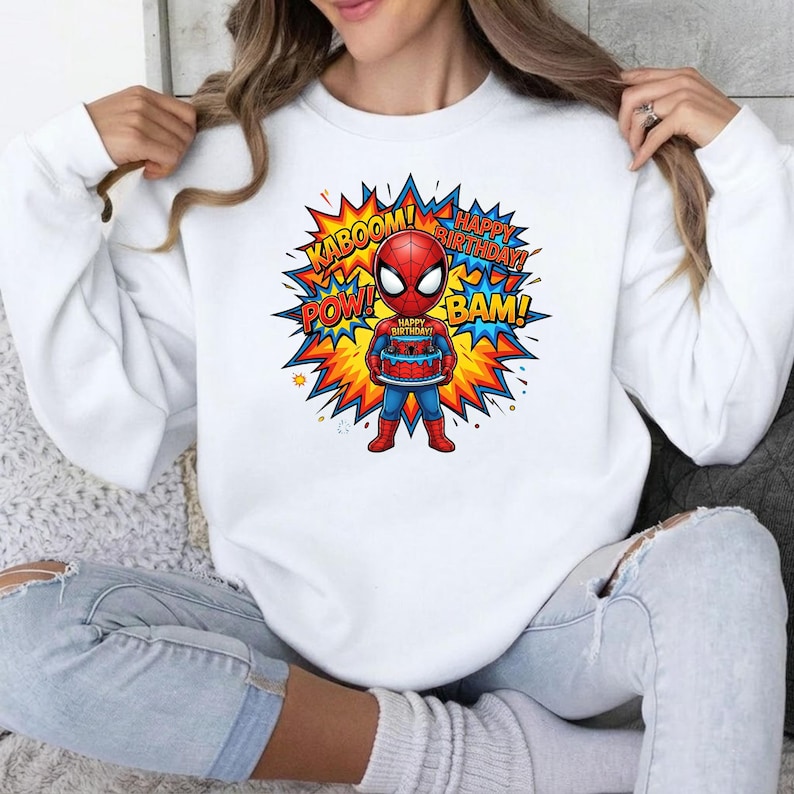 May include: A white long-sleeve sweatshirt featuring a graphic of a cartoon Spider-Man holding a birthday cake. The design includes colorful comic book style bursts with the text KABOOM, POW, BAM, and HAPPY BIRTHDAY in bold, vibrant lettering.