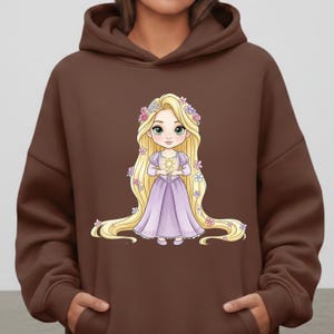 Op de afbeelding: Een bruine hoodie met een cartoonillustratie van een prinses met lang blond haar, gekleed in een lavendelkleurige jurk en met een gouden zon. De prinses is versierd met bloemmotieven.