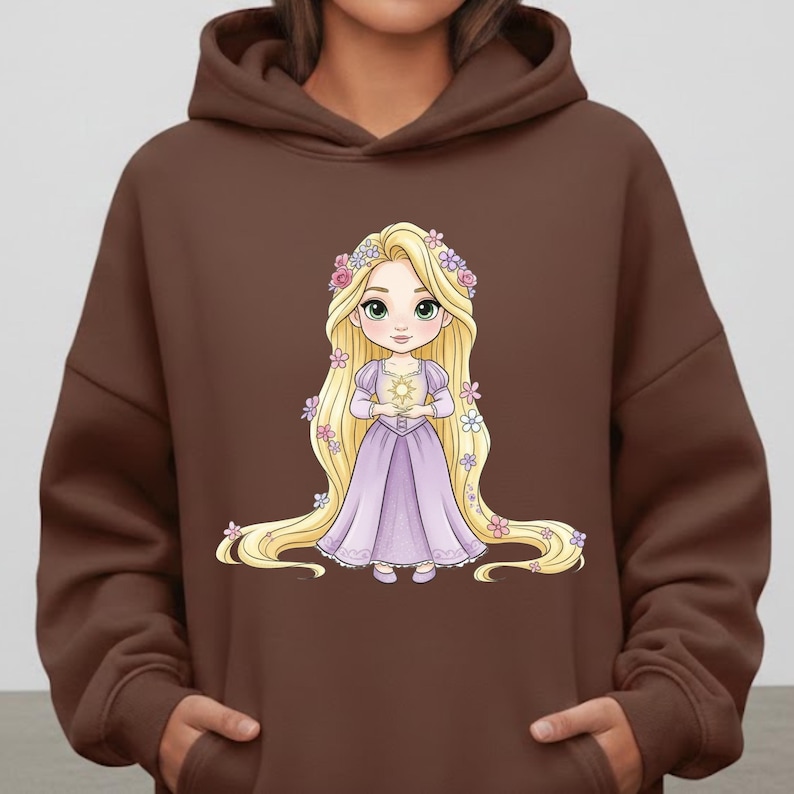 Op de afbeelding: Een bruine hoodie met een cartoonillustratie van een prinses met lang blond haar, gekleed in een lavendelkleurige jurk en met een gouden zon. De prinses is versierd met bloemmotieven.