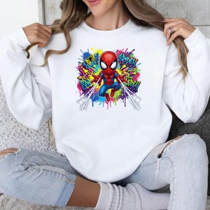 May include: A white long-sleeve sweatshirt featuring a central graphic of a cartoon Spider-Man character in red and blue. The design includes colorful paint splatters in yellow, purple, and green, with the text Spidey written in graffiti style.