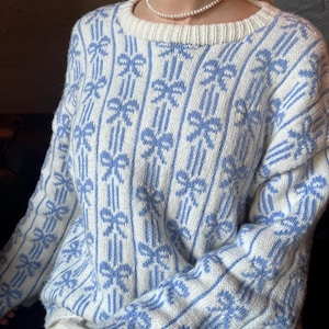 Könnte beinhalten: Ein weiß-hellblauer Pullover mit Schleifenmuster. Der Pullover hat einen Rundhalsausschnitt und lange Ärmel. Die Schleifen sind hellblau und in vertikalen Streifen angeordnet. Der Pullover ist aus einem weichen Strickmaterial gefertigt.