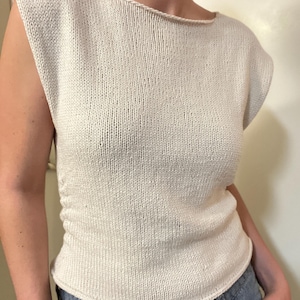 Puede incluir: Una camiseta de punto sin mangas de color crema con cuello barco. La parte superior tiene un aspecto ligeramente texturizado y una silueta ajustada. El dobladillo se curva suavemente. La parte superior está hecha de un hilo suave y claro.