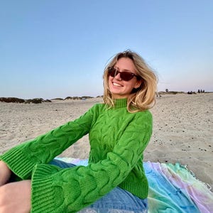 May include: A bright green cable-knit sweater with flared sleeves. The sweater is paired with blue jeans and sunglasses. The person is sitting on a colorful tie-dye blanket on a sandy beach under a clear sky.