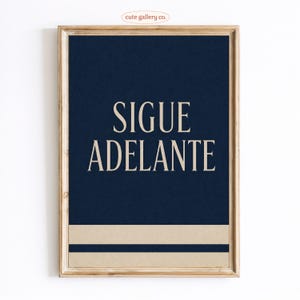 May include: Framed art print with a navy blue background and the words "SIGUE ADELANTE" in a cream-colored font. A cream-colored horizontal stripe is at the bottom. The frame is a light brown color. The text is in Spanish.