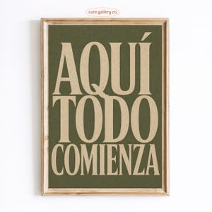 May include: A framed print with the text "AQUÍ TODO COMIENZA" in large, beige letters on a dark green background. The frame is a light brown color. The words translate to "Here everything begins". The words "cute gallery co." are in the top left corner.