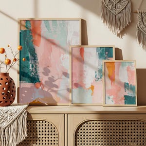 Könnte beinhalten: Vier gerahmte abstrakte Kunstdrucke mit moderner Ästhetik. Das Kunstwerk zeigt Pinselstriche in Rosa, Türkis, Orange und Weiß. Die Rahmen sind aus hellem Holz und die Drucke werden auf einem Holzschrank ausgestellt.