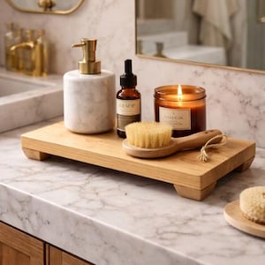 May include: A wooden tray holds a marble soap dispenser with a gold pump, a brown bottle, a body brush, and a lit candle. The items are arranged on a marble countertop in a bathroom setting. The candle has a label that reads "Sallow".