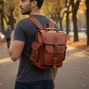 Peut inclure: Un sac à dos en cuir marron avec plusieurs poches et une poignée supérieure. Le sac à dos a des bretelles réglables et une fermeture à boucle. La personne porte un t-shirt gris et un jean bleu.