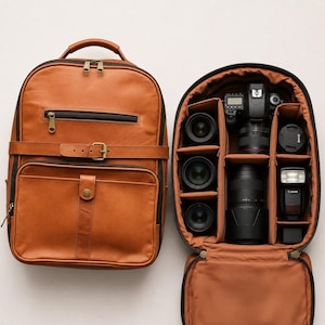 May include: A tan leather camera backpack with a zippered front pocket and a horizontal strap. Beside it, the open interior reveals a tan lining with padded compartments containing a black camera body, three lenses, a flash unit, and accessories.