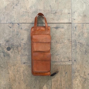 Peut inclure: Un sac en cuir marron vertical avec une poignée et trois poches avant. Le sac est rectangulaire avec des coins arrondis et une tirette de fermeture éclair. Le cuir a un aspect naturel et texturé.