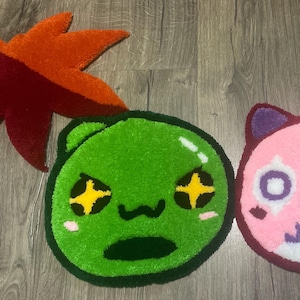 May include: Two tufted rugs shaped like cartoon characters, one green with star-shaped eyes and one pink with purple ears, accompanied by two leaf-shaped rugs in orange and dark red, all featuring a textured, plush pile surface.