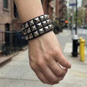 Puede incluir: Pulsera de cuero negro con tres filas de tachuelas piramidales plateadas. La pulsera es un accesorio audaz, perfecto para añadir un toque atrevido a cualquier atuendo. Las tachuelas reflejan la luz.