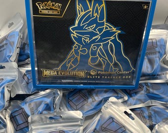 Puede incluir: Una vitrina de acrílico azul que contiene una caja de entrenador de élite Pokémon Center Mega Evolution con un gráfico de Lucario. La caja presenta el logotipo y el texto del juego de cartas coleccionables Pokémon. Varias bolsas de plástico pequeñas y selladas que contienen protectores de bordes de silicona azul están dispuestas en primer plano.