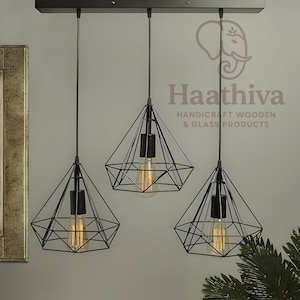 Può includere: Tre lampade a sospensione geometriche nere con lampadine a vista sono appese a un apparecchio rettangolare. Le luci hanno un design a gabbia a forma di diamante. L'immagine include il testo "Haathiva HANDICRAFT WOODEN & GLASS PRODUCTS".