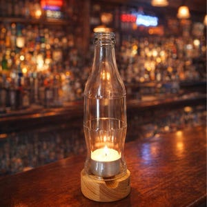 May include: A clear glass bottle-shaped candle holder with a lit tea light inside, set on a wooden base. The candle holder is on a dark wooden surface, with a blurred bar background.