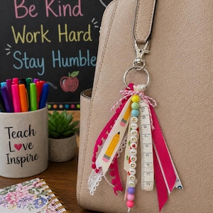 May include: A bag charm featuring a silver lobster clasp and ring, adorned with various ribbons in pink and white, a pencil-patterned ribbon, a measuring tape ribbon, and a strand of colorful beads spelling MISS with a heart and apple icons.
