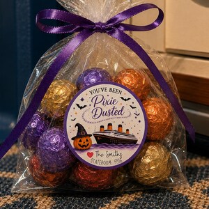 May include: A clear cellophane gift bag filled with round candies wrapped in purple, orange, and gold foil. The bag is tied with a purple ribbon and features a circular sticker that reads: You've Been Pixie Dusted, The Smiths Stateroom 9516.