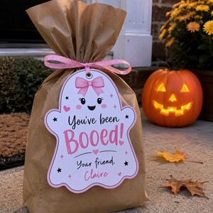 May include: A brown paper gift bag tied with a pink polka dot ribbon and a white ghost-shaped tag. The tag features a pink bow, black eyes, a smiling mouth, and the text "You've been Booed! Your friend, Claire" in pink and black lettering.
