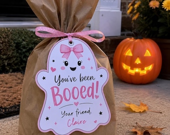 May include: A brown paper gift bag tied with a pink polka dot ribbon and a white ghost-shaped tag. The tag features a pink bow, black eyes, a smiling mouth, and the text "You've been Booed! Your friend, Claire" in pink and black lettering.