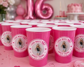 May include: A collection of bright pink plastic party cups, each featuring a circular sticker with a photograph of a person, the text "LOOK WHO'S 16!", the name "GRACE YOUNG", and "EST. 2010" surrounded by small heart and star icons.