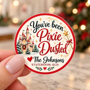 May include: A round sticker with a red border featuring a castle, a gold star wand, and the text "You've been Pixie Dusted", "The Johnsons", and "STATEROOM: 8124". The design includes green pine trees, red hearts, and gold sparkles on a cream background.