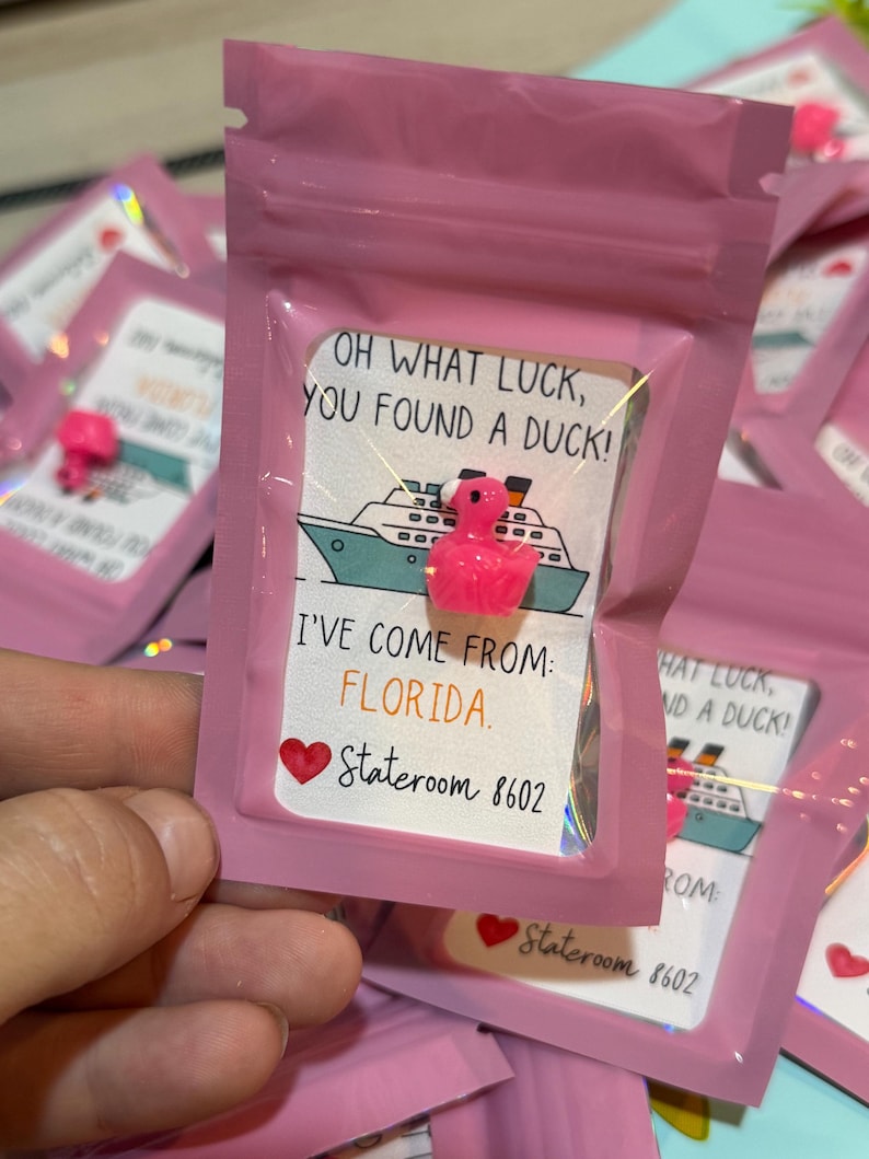 Puede incluir: Una peque&ntilde;a figura de pato de pl&aacute;stico rosa dentro de una bolsa rosa resellable. El empaque incluye una tarjeta con una ilustraci&oacute;n de un crucero y el texto: OH WHAT LUCK, YOU FOUND A DUCK! I'VE COME FROM: FLORIDA. Un coraz&oacute;n rojo y Stateroom 8602.