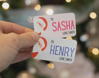 May include: Two rectangular white gift tags featuring a cartoon Santa Claus face on the left side. The top tag reads TO: SASHA LOVE SANTA in pink text. The bottom tag reads TO: HENRY LOVE SANTA in blue text with black accents.