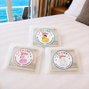 May include: Three translucent envelopes containing folded US currency, each sealed with a circular sticker. The stickers feature the text THANK YOU, The Field Family, Stateroom 1234, and illustrations of a yellow duck, a pink flamingo, or an elephant.