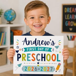May include: A white rectangular sign featuring colourful text and graphics. The text reads Andrew's FIRST DAY OF PRESCHOOL 2026-2027. The design includes illustrations of a pencil, paper aeroplanes, stars, and hearts in blue, green, orange, and yellow.