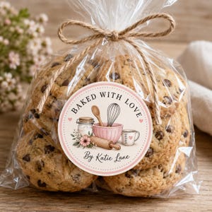 May include: A clear plastic bag tied with twine containing chocolate chip cookies. A round sticker on the front features a pink bowl, whisk, wooden spoon, rolling pin, and measuring cup with the text Baked With Love By Katie Lane.