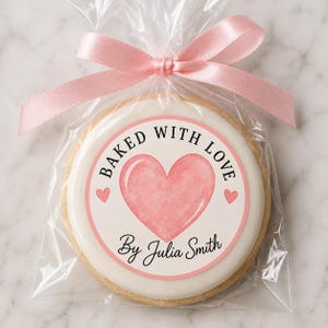 May include: A round cookie with white icing and a pink heart graphic, featuring the text "BAKED WITH LOVE" and "By Julia Smith" in black, enclosed in a clear plastic bag tied with a light pink satin ribbon at the top of the packaging.