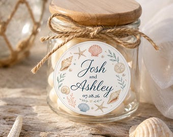 May include: A clear glass jar with a wooden lid, tied with a jute twine bow. The front label features a circular design with watercolor seashells, starfish, and greenery, displaying the text Josh and Ashley with the date 07.28.26.