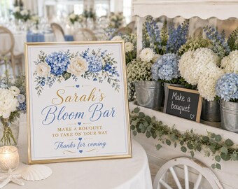 May include: A rectangular sign in a gold frame featuring a floral illustration of blue hydrangeas and white roses. The text reads: Sarah's Bloom Bar, MAKE A BOUQUET TO TAKE ON YOUR WAY, and Thanks for coming, accented with small blue hearts.