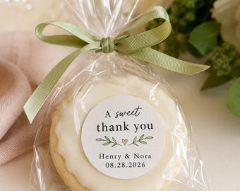 May include: A round cookie with white icing inside a clear plastic bag tied with a sage green ribbon. A circular sticker on the bag reads A sweet thank you, Henry & Nora, 08.28.2026, with a small heart and leaf illustrations in the center.