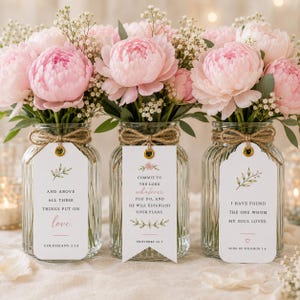 May include: Three clear ribbed glass jars filled with light pink peonies and baby's breath. Each jar features a jute twine bow and a white rectangular tag with floral illustrations and printed Bible verses: Colossians 3:14, Proverbs 16:3, and Song of Solomon 3:4.