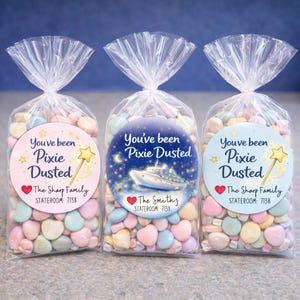 May include: Three clear cellophane bags filled with pastel-colored candies. Each bag is sealed with a twist tie and a circular sticker that reads "You've been Pixie Dusted." The stickers feature a cruise ship, a wand, and family names.