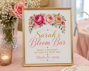May include: A rectangular gold-toned frame containing a white sign with a floral illustration of pink and peach peonies at the top. The text reads: Sarah's Bloom Bar, MAKE A BOUQUET TO TAKE ON YOUR WAY, Thanks for coming, accented with small red hearts.