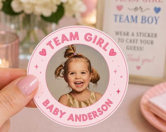 May include: A circular pink sticker featuring a photograph of a smiling child with pigtails in the center. The text reads TEAM GIRL at the top and BABY ANDERSON at the bottom, accented with pink hearts and small star icons on the border.