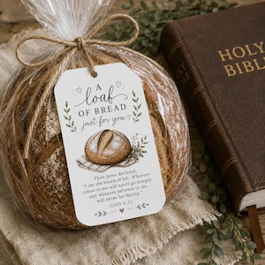 Op de afbeelding: Een rond brood in een doorzichtige cellofaan zak, vastgebonden met touw, en een cadeaulabel met de tekst "Een brood alleen voor jou". Een bruine, ledergebonden bijbel is zichtbaar op de achtergrond. Het label bevat een bijbelvers en een tekening van een brood.