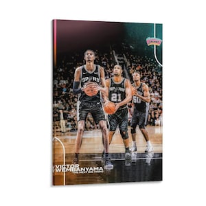 May include: A canvas print featuring three basketball players in black and white uniforms, holding basketballs. The players are on a court with a crowd in the background. The text "VICTOR WEMBANYAMA THE NEXT BIG THING" is at the bottom.
