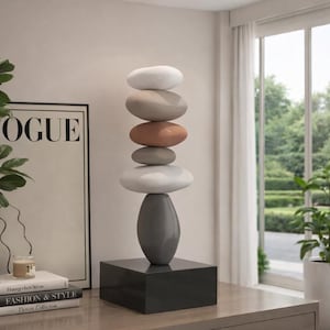 May include: A modern sculpture featuring stacked, smooth, oval-shaped stones in shades of white, tan, and terracotta, balanced atop a gray, teardrop-shaped base. The base sits on a dark gray square pedestal. The sculpture is a decorative accent piece.