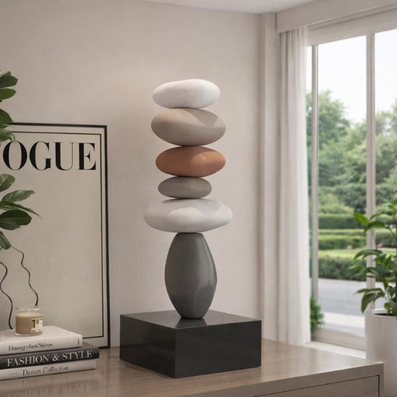 May include: A modern sculpture featuring stacked, smooth, oval-shaped stones in shades of white, tan, and terracotta, balanced atop a gray, teardrop-shaped base. The base sits on a dark gray square pedestal. The sculpture is a decorative accent piece.
