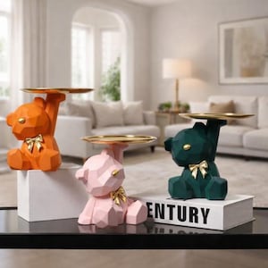 May include: Three decorative bear figurines in orange, pink, and dark green, each holding a gold-coloured tray. The bears have geometric designs and gold bow accents. The figurines are displayed on a black table.
