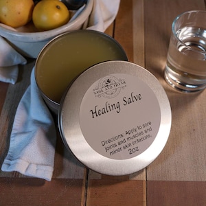 May include: A round metal tin containing a pale yellow healing salve. The lid features a white label with the text Sage and Thyme Healing Salve, directions for use on sore joints and muscles, and the weight of 2oz printed in black ink.