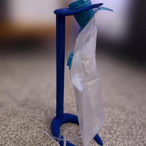 May include: A blue plastic stand with a vertical support and a circular base, holding a translucent white medical drainage bag. The bag features a blue cap at the top and a clear plastic tube extending from the bottom, hanging vertically from the stand.